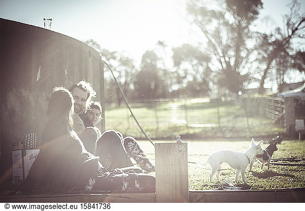 Friends with dogs sit and laugh outside in sunlight on farm