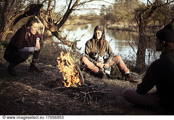 Friends with dogs sit around campfire by river in spring in germany