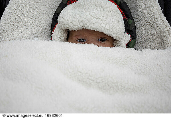 From above of funny cute little baby in warm hat covered with fur looking at camera while lying in stroller in wintertime