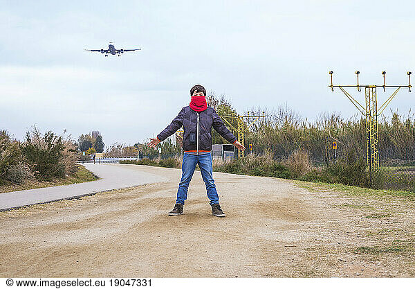 Front view of a boy standing with outstretched arms and wearing a red scarf while an airplane taking off in airport