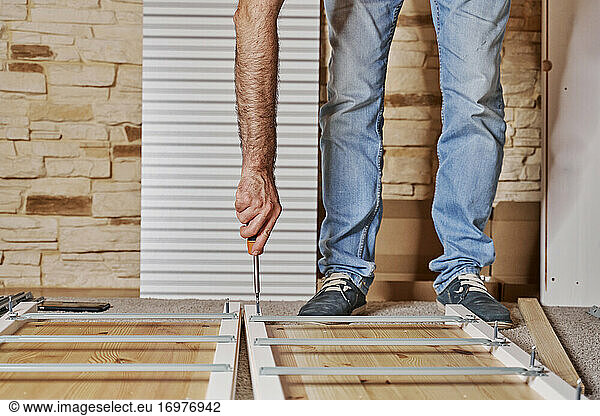 Front view of a unrecognized worker screwing a screw with a screwdriver in a strip of a furniture assembling by himself a wardrobe of wood. Horizontal photo.