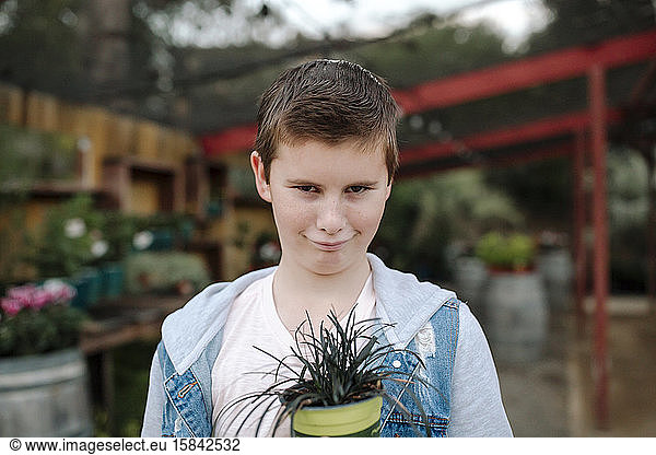 Frowning preteen boy in denim jacket holding grassy plant at nursery