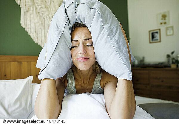 Frustrated woman covering ears with pillow in bed at home