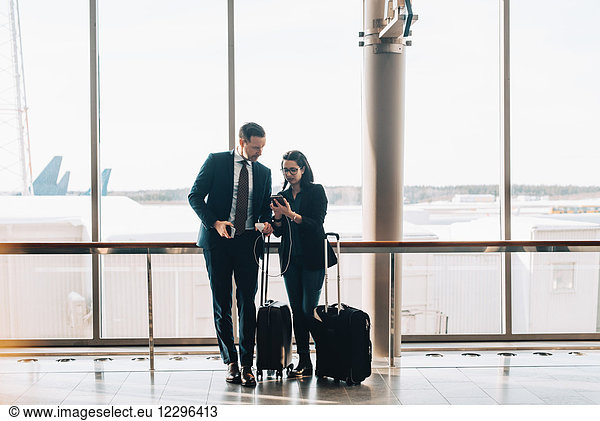 Full length of business couple with luggage using mobile phone in airport terminal
