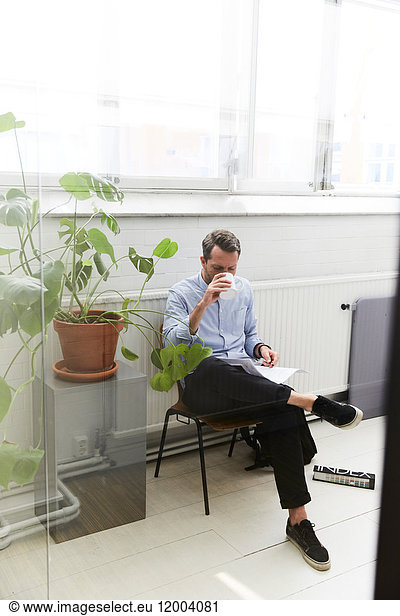 Full length of businessman having coffee while reading document by potted plant at office