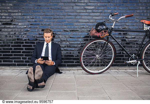 Full length of businessman using digital tablet while sitting on sidewalk by bicycle
