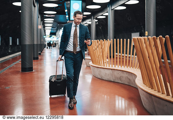 Full length of businessman with luggage and mobile phone walking in railroad platform
