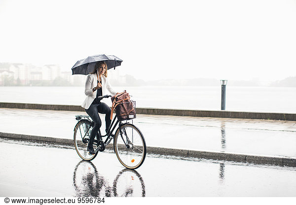Full length of businesswoman riding bicycle on wet city street during rainy season
