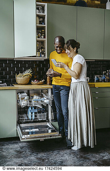 Full length of cheerful woman showing smart phone to boyfriend doing dishes in kitchen at home