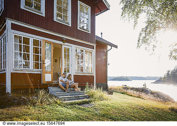 Full length of couple sitting at log cabin entrance by lake during summer