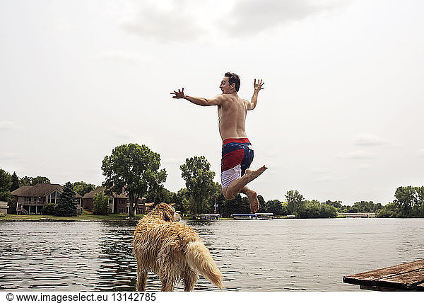 Full length of excited man with dog jumping in lake against sky