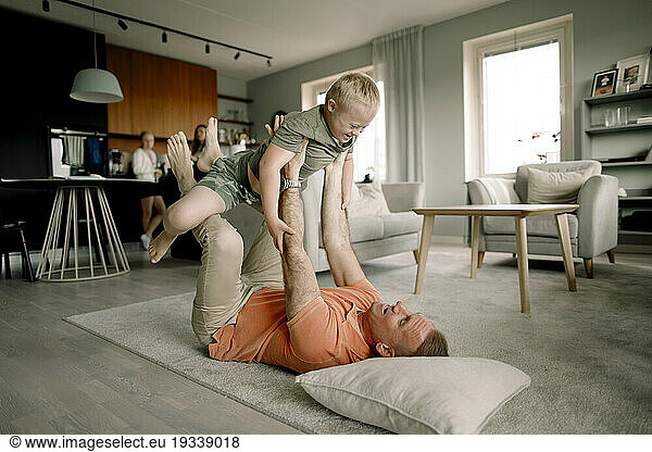 Full length of father lying on carpet while lifting son in living room at home