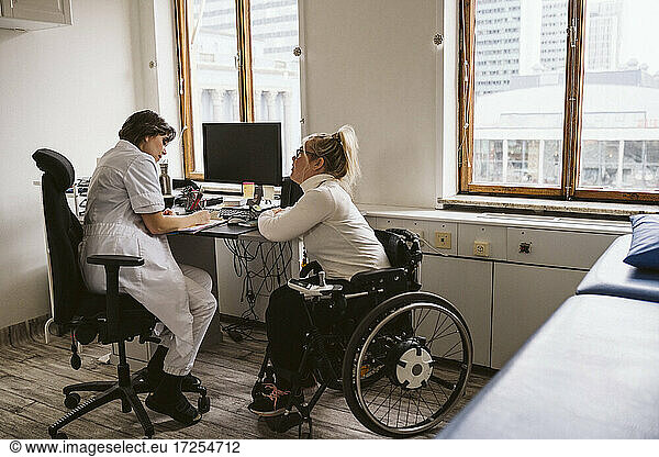Full length of female doctor writing prescription to disabled patient while sitting at desk