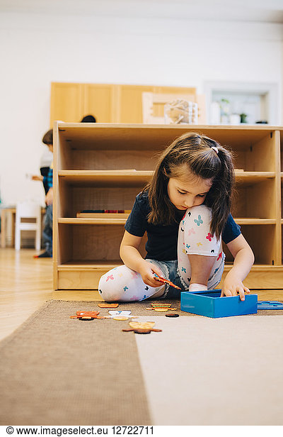 Full length of girl playing with toys while sitting on floor against rack in child care classroom