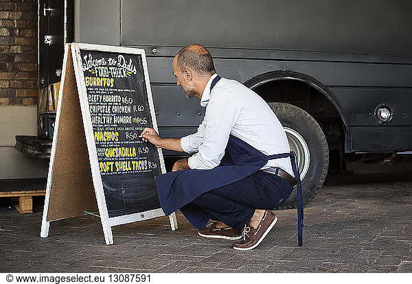 Full length of male vendor writing on blackboard while crouching at street by food truck