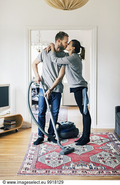 Full length of man kissing woman while vacuuming carpet at home