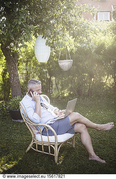 Full length of man talking on smart phone while using laptop in backyard