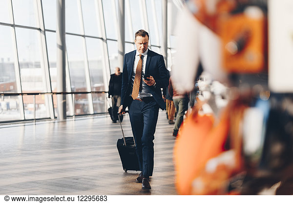 Full length of mature businessman with luggage using mobile phone while walking in airport terminal