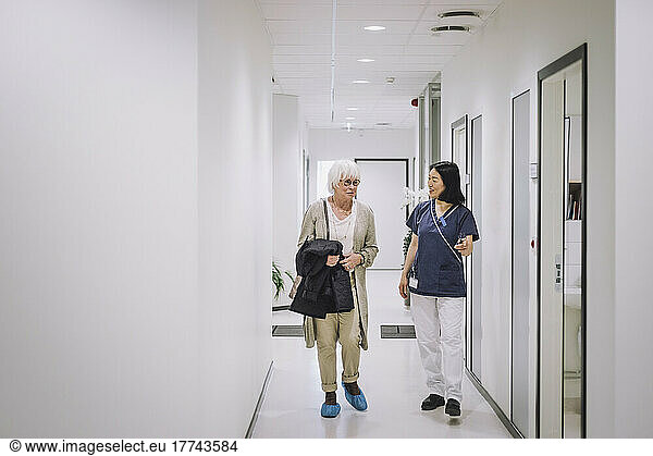 Full length of senior woman talking with female doctor while walking in corridor at medical clinic