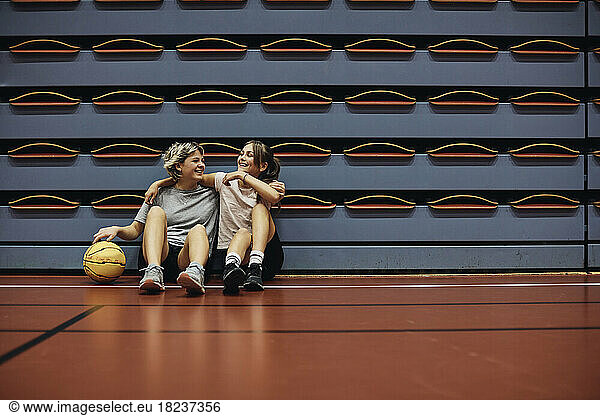Full length of smiling girls sitting with arms around each other at basketball court in school