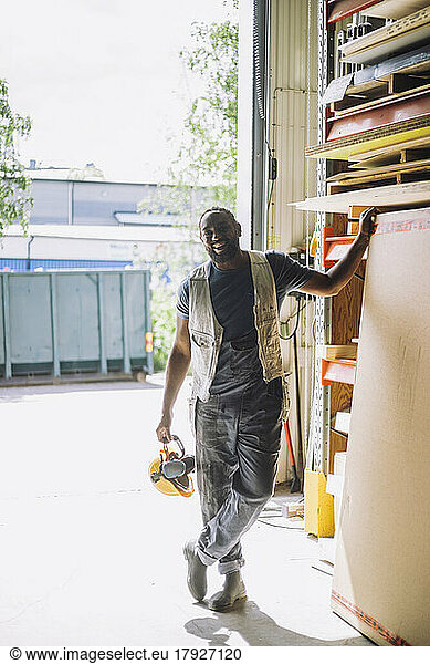 Full length of smiling male carpenter with hardhat standing by wooden plank in warehouse