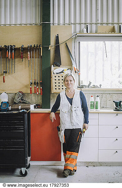 Full length of smiling mature female carpenter standing by workbench in workshop