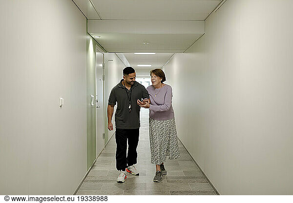 Full length of smiling senior woman walking arm in arm with male nurse at retirement home corridor