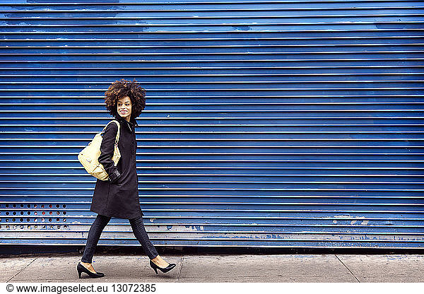 Full length of smiling woman walking on sidewalk by blue shutter