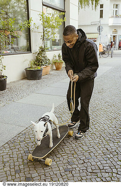Full length of smiling young man walking with Bull Terrier on skateboard at sidewalk