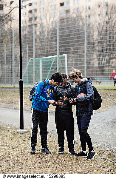 Full length of social media addicted friends wearing sports clothing while using mobile phones on playing field