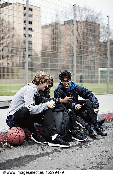 Full length of social media addicted friends wearing warm clothing while sitting on sidewalk after basketball practice d