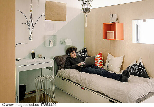 Full length of teenage boy using laptop while lying on bed at home
