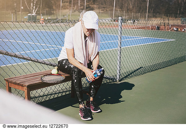 Full length of tired woman sitting on bench against net at tennis court