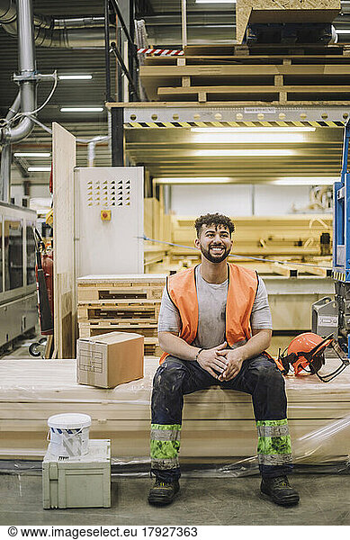 Full length of young carpenter sitting by hardhat in warehouse while looking away