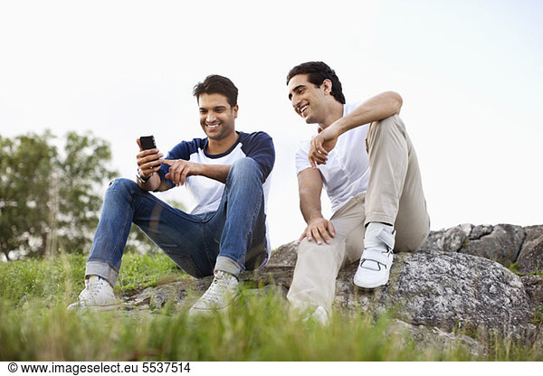 Full length of young friends sitting on rock with mobile phone