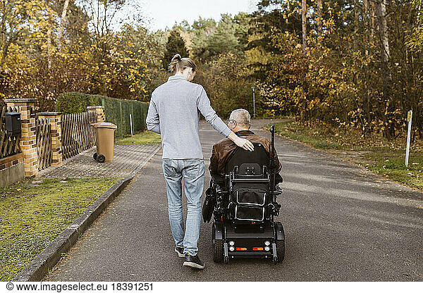 Full length of young male caregiver walking by senior man with disability in motorized wheelchair on road
