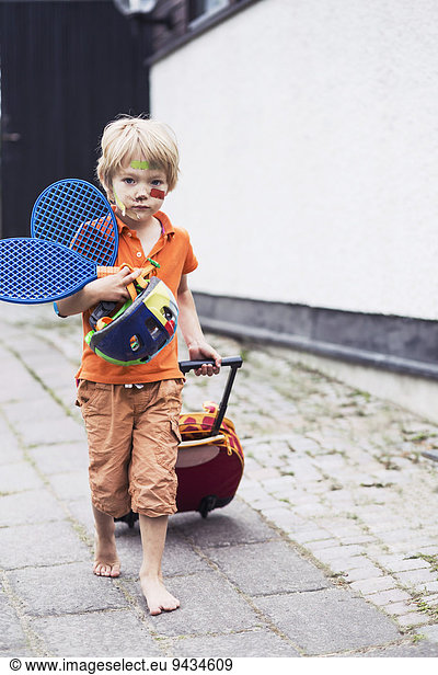Full length portrait of boy pulling luggage while holding tennis rackets and helmet on street