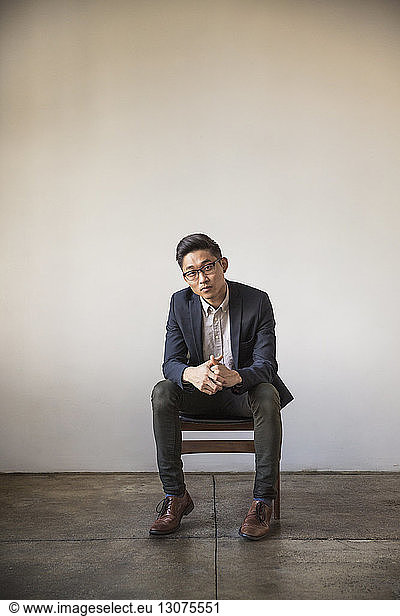 Full length portrait of confident businessman sitting on chair against wall in office