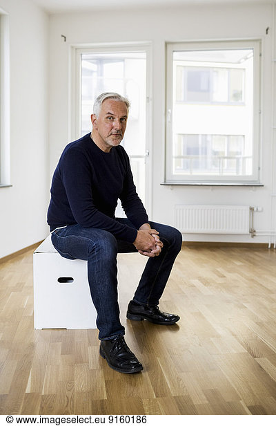Full length portrait of confident man sitting on moving box in new house