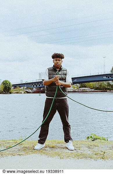 Full length portrait of confident young man holding garden hose near river