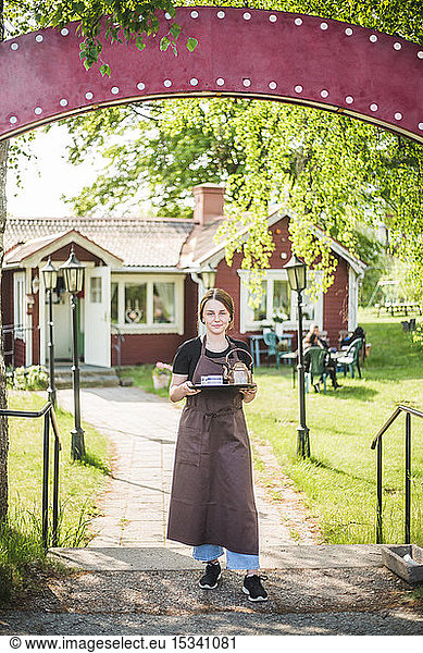 Full length portrait of confident young waitress holding serving tray while standing at outdoor cafe entrance
