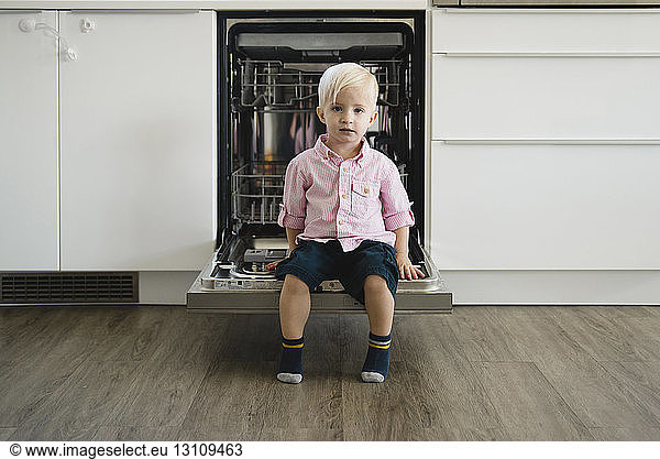 Full length portrait of cute boy sitting on dishwasher door at home