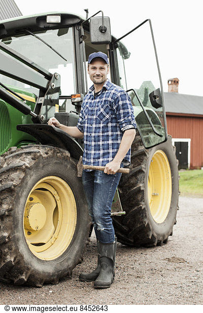 Full length portrait of farmer with hammer standing by tractor on farm