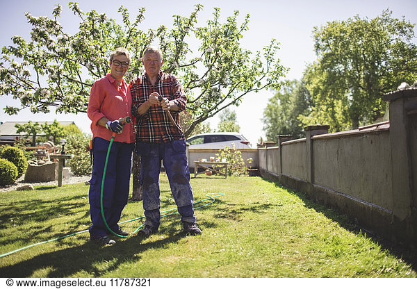 Full length portrait of senior couple holding hammer and garden hose while standing at yard