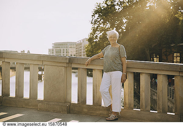 Full length portrait of senior woman standing on bridge against railing in city