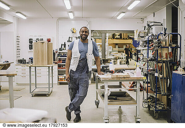 Full length portrait of smiling carpenter standing on one leg while leaning on workbench at workshop