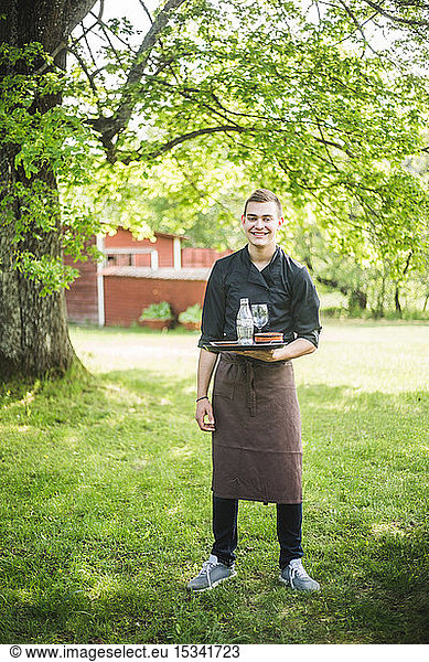 Full length portrait of smiling confident young waiter standing on grass at outdoor cafe