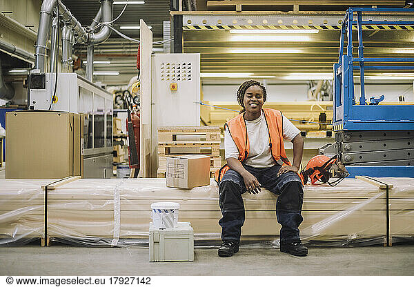 Full length portrait of smiling female carpenter sitting by hardhat in warehouse