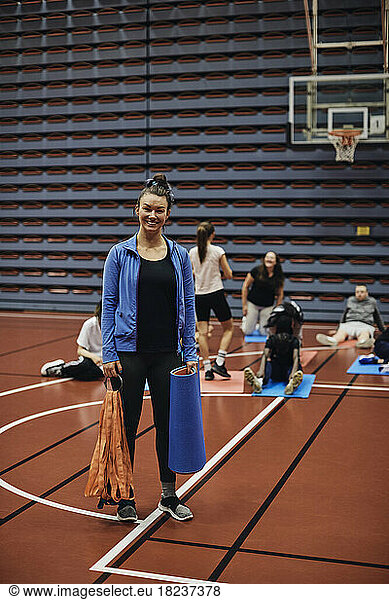 Full length portrait of smiling female coach standing with exercise mat in sports court