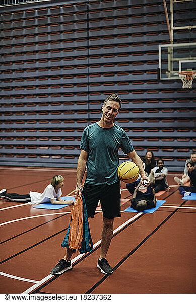 Full length portrait of smiling male coach standing with basketball against students at sports court